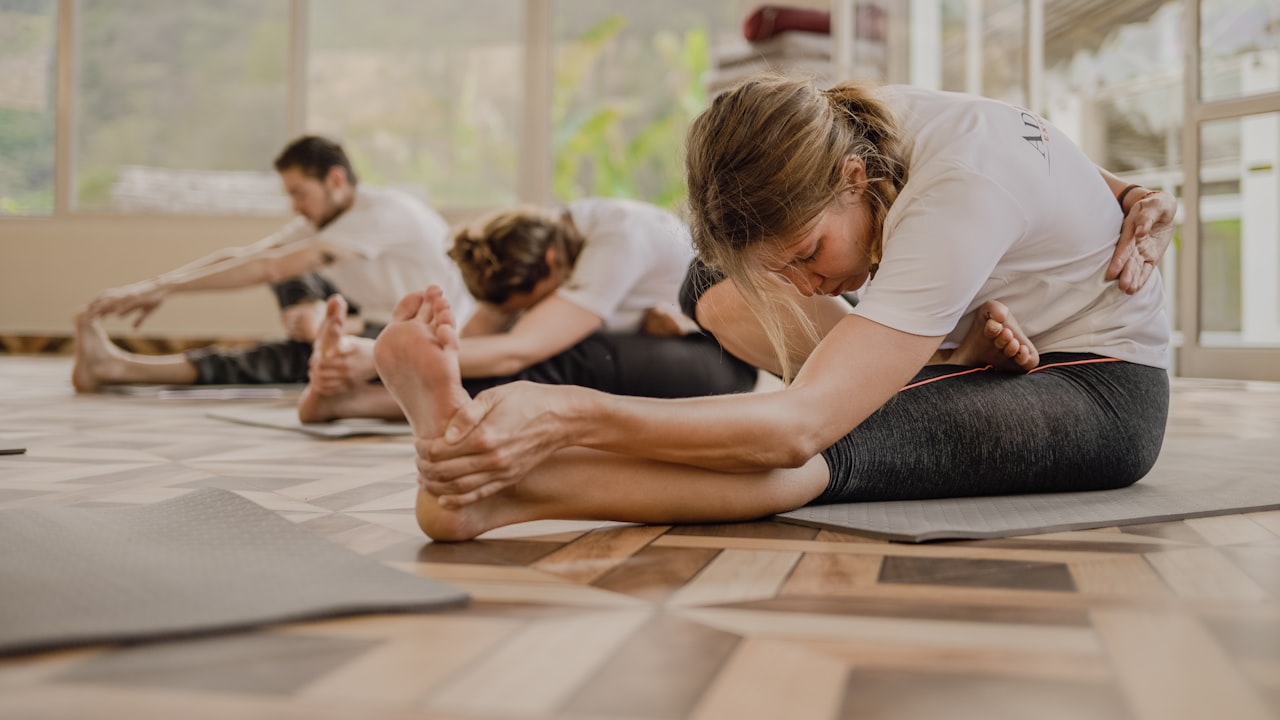 Un groupe de quatre personnes assises sur des tapis de sol, en pleine étirement avant lors d'un cours de yoga collectif dans une salle lumineuse