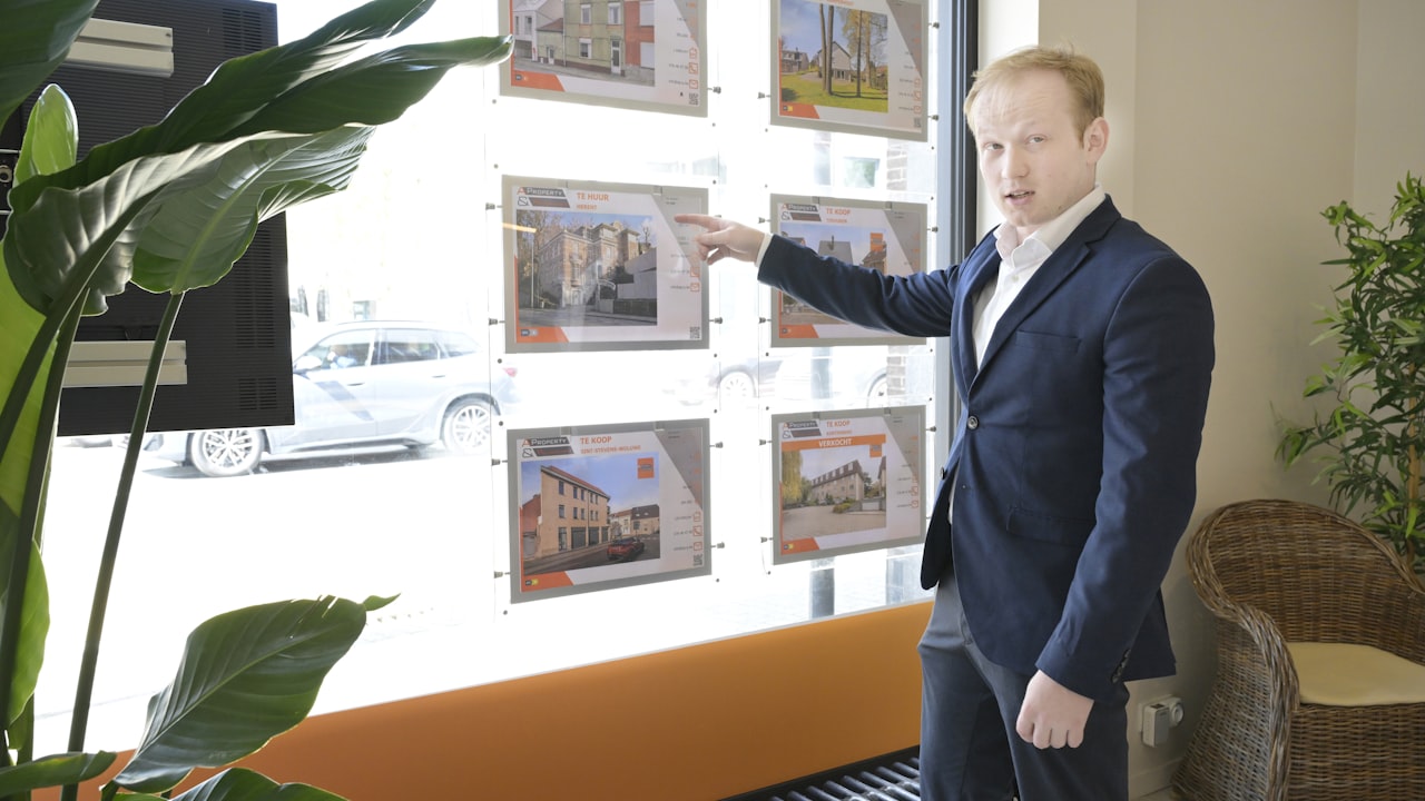Un négociateur immobilier en costume montre du doigt les annonces de biens affichées dans la vitrine de son agence, devant des plantes vertes
