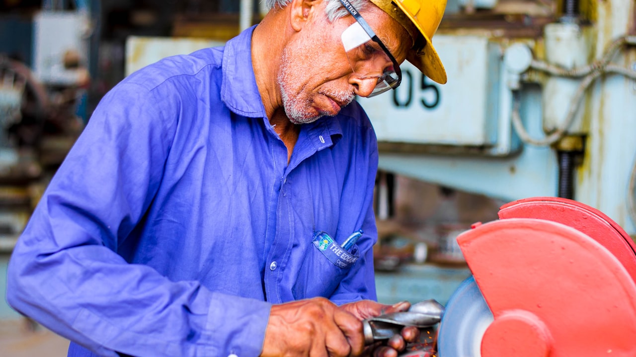 Un opérateur âgé, casque de chantier jaune et lunettes de protection, meule une pièce métallique à mains nues sur un touret à meuler, des étincelles jaillissant dans un atelier industriel
